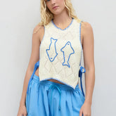 Woman wearing a white sleeveless top with blue fish designs and blue pants on a plain background